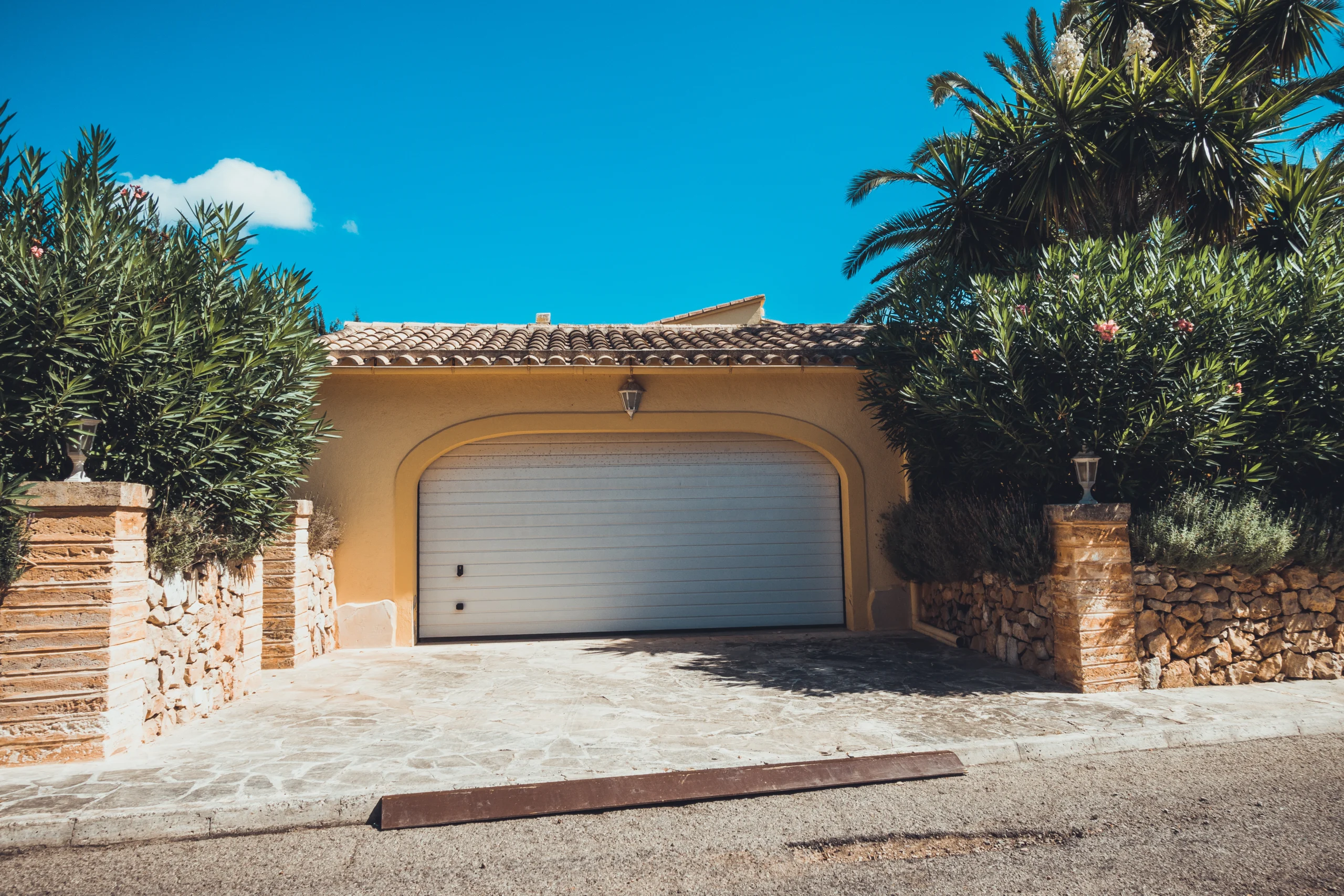 A yellow house with a white garage door and lush green foliage.