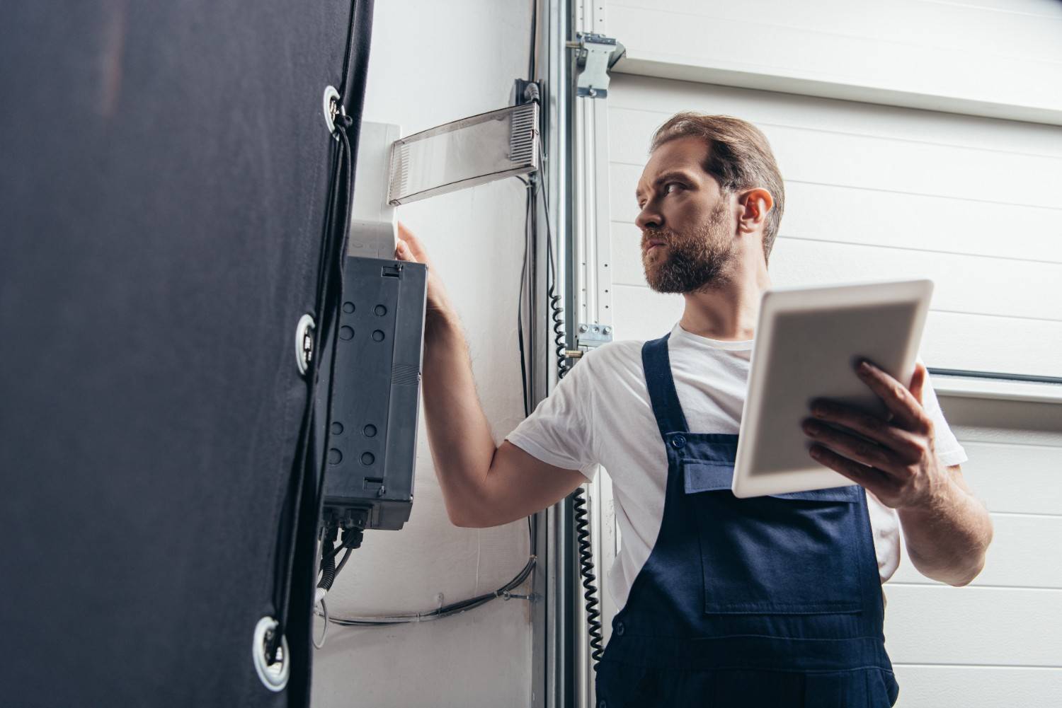 Man in overalls adjusting a control panel while holding a tablet.
