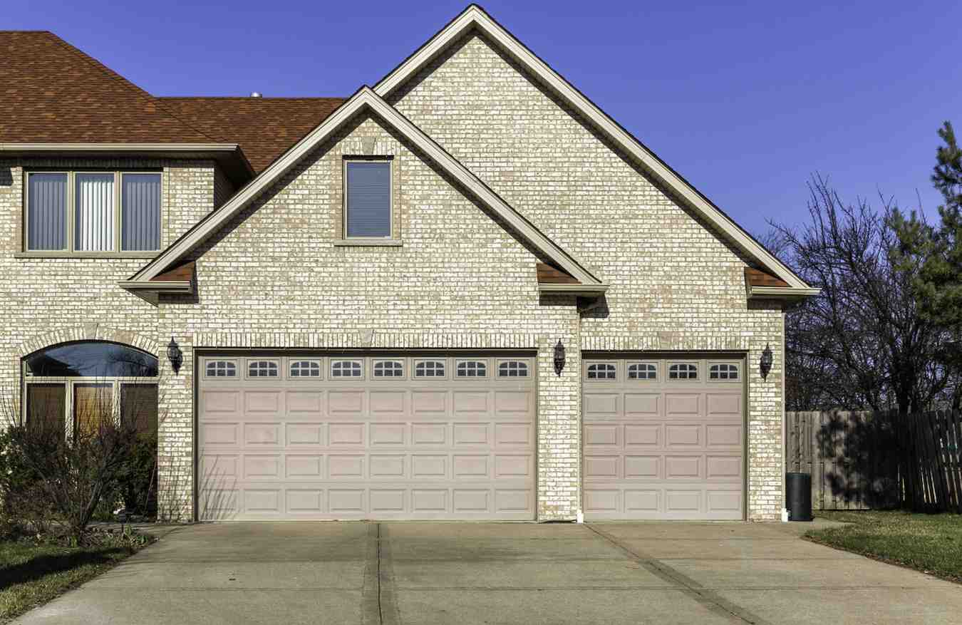 Two beige garage doors on a light brick house under a blue sky.